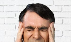 A man in front of a white brick wall holding his temples on his face with his hands and scrunching his face with his eyes closed.