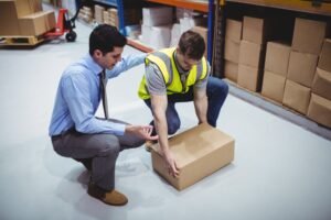 A worker inside a warehouse attempts to lift a heavy box with the assistance of a colleague