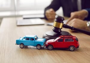 A solicitor works on a road traffic accident claim in the background. There are two small crashed toy cars on the desk in the foreground.