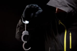 A hooded and masked person with handcuffs dangling from one hand. 