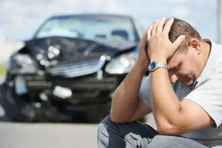 A man with his head in his hands and his car is damaged in the background.