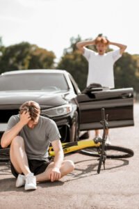 The aftermath of a cycling and car accident with the bike under the wheels, cyclist sat in front of the car and driver behind the open door. 