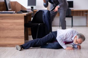 An office worker lying on the floor in pain next to his desk while his colleague is on the phone frantically calling for help.