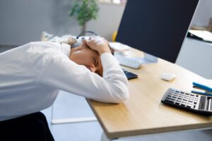 An office worker lying down at her desk holding her head in pain next to a computer, stapler and calculator.
