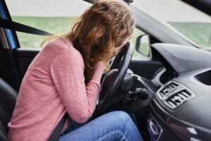 A woman sitting in her car with her face in her hands leaning against the steering wheel after being involved in a hit and run.